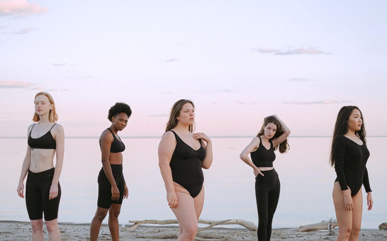 Five women of diverse backgrounds posing confidently on a serene beach.